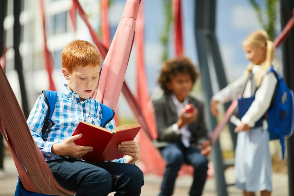 Un écolier assis lit un livre de couverture rouge devant d'autres enfants.