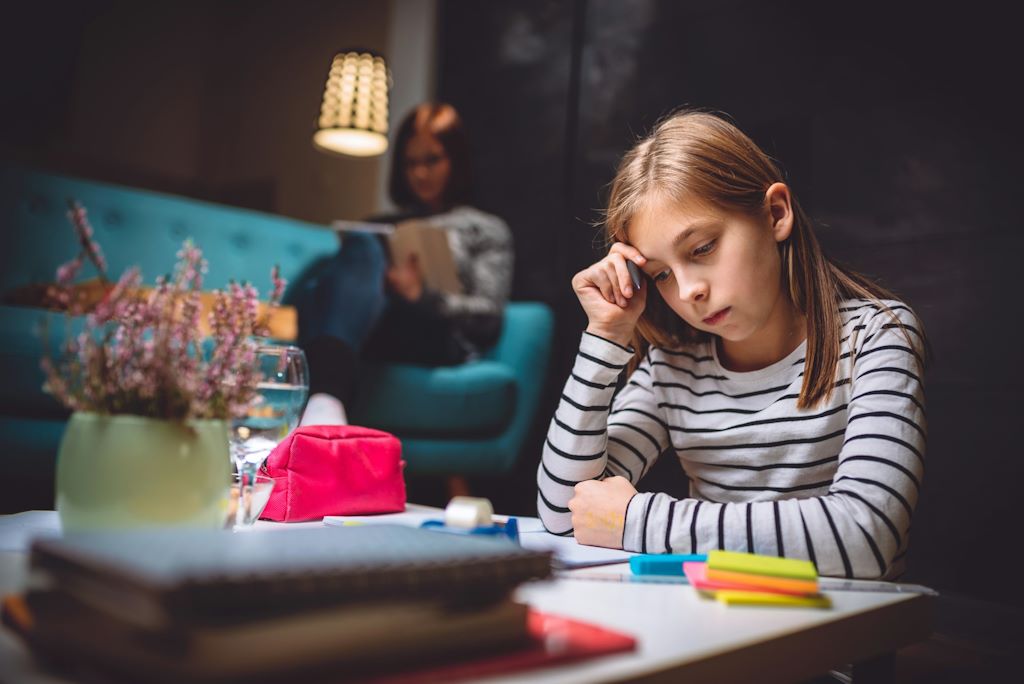 Une écolière concentrée sur ses devoirs, devant un canapé où se repose une adulte.