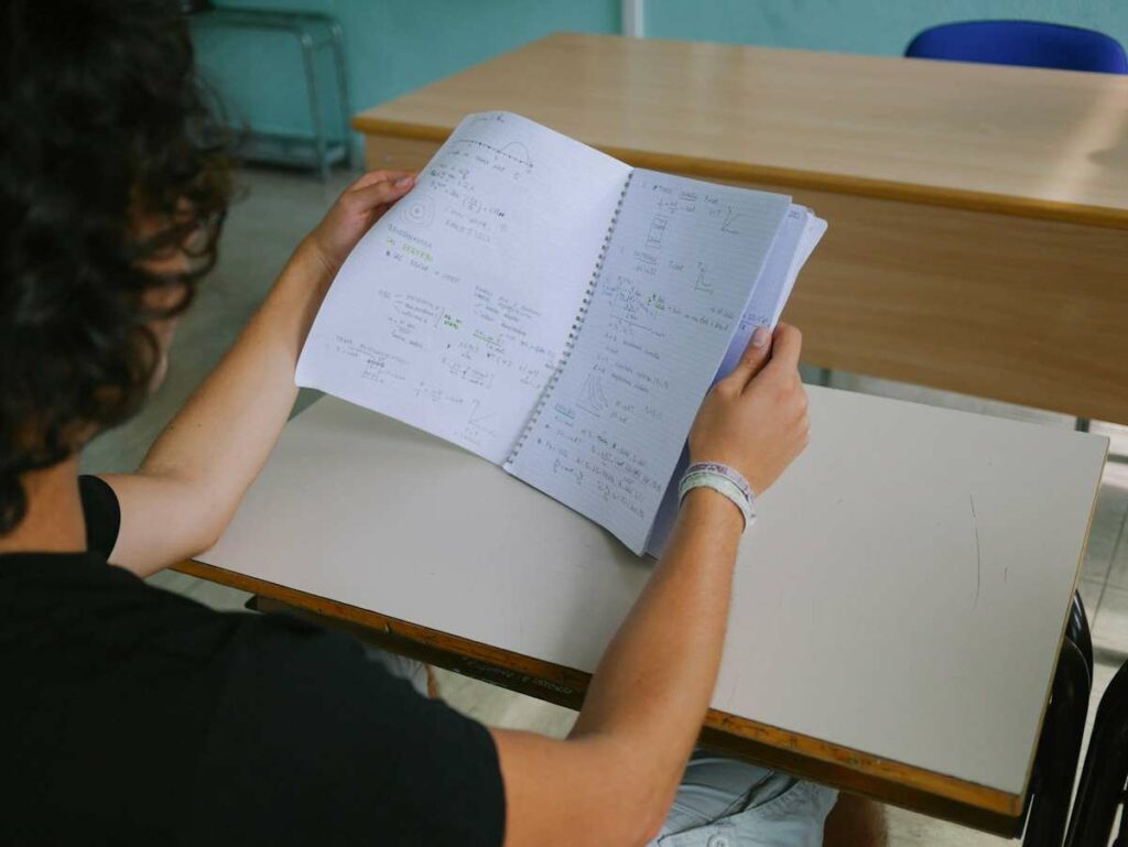 Un élève tient ouvert un cahier avec des exercices de mathématiques, des formules et des calculs.