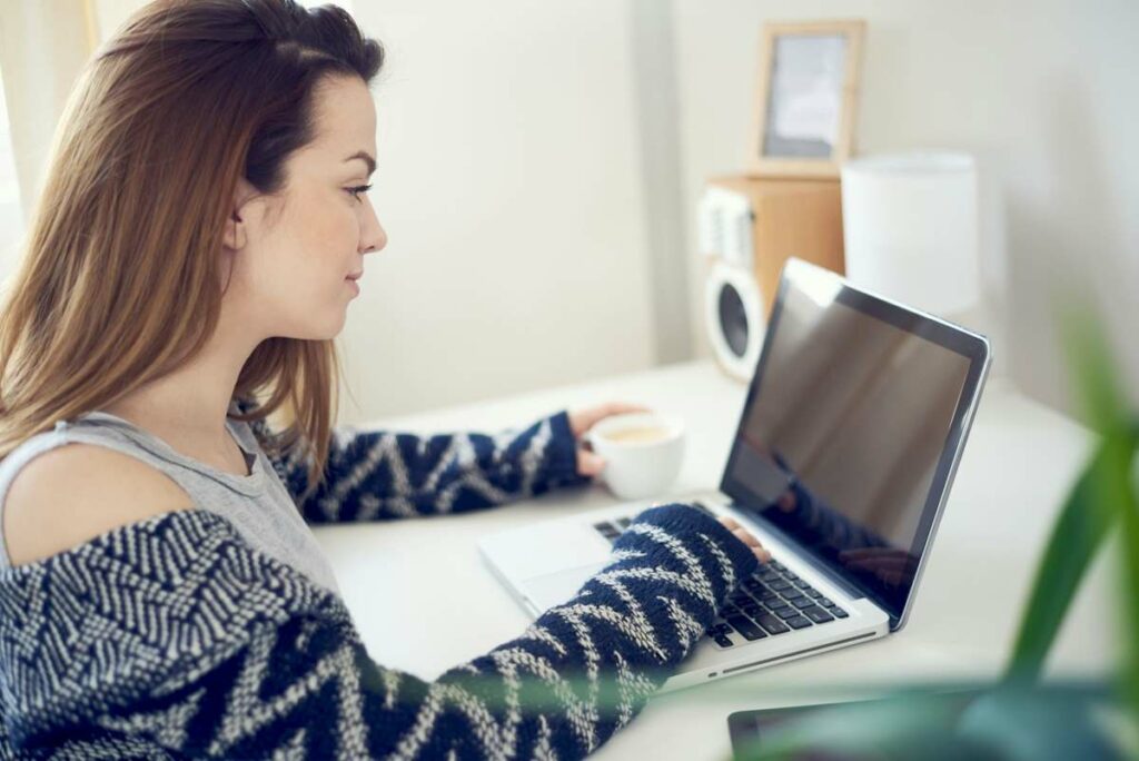Une étudiante travaille devant son écran en tenant une tasse de café de sa main gauche.