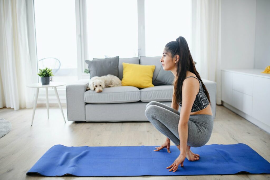 Une femme fait une séance de yoga sur un tapis bleu, dans une pièce lumineuse.