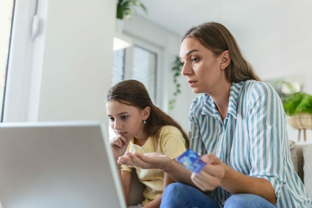 Une mère de famille avec sa fille devant un écran, tient une carte bancaire de sa main gauche.