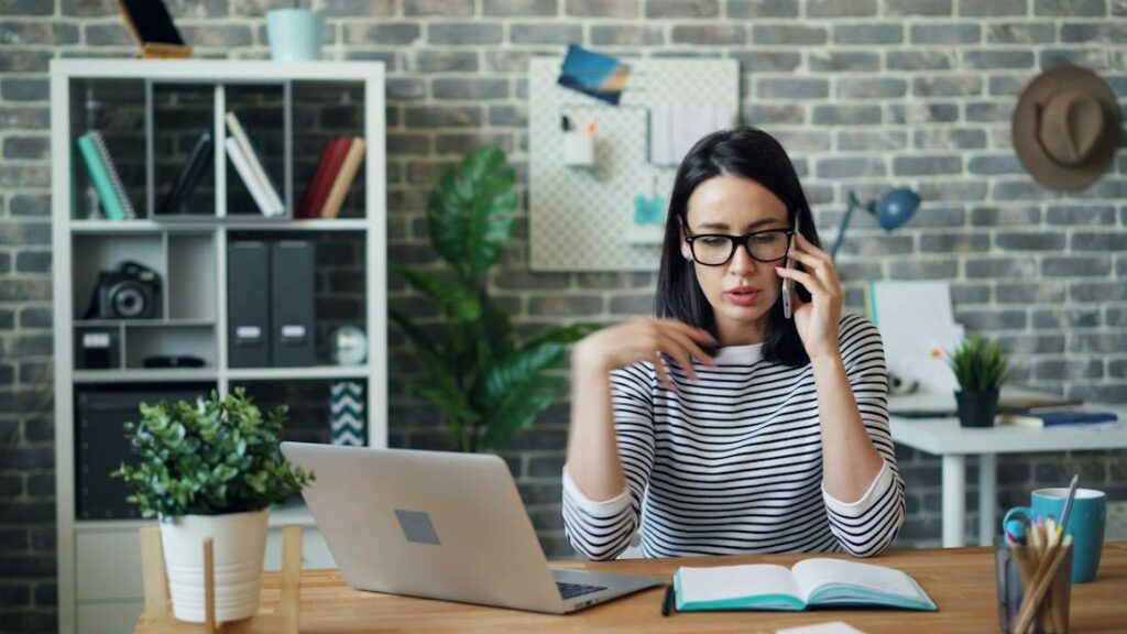 Une femme parle avec son smartphone devant un ordinateur.