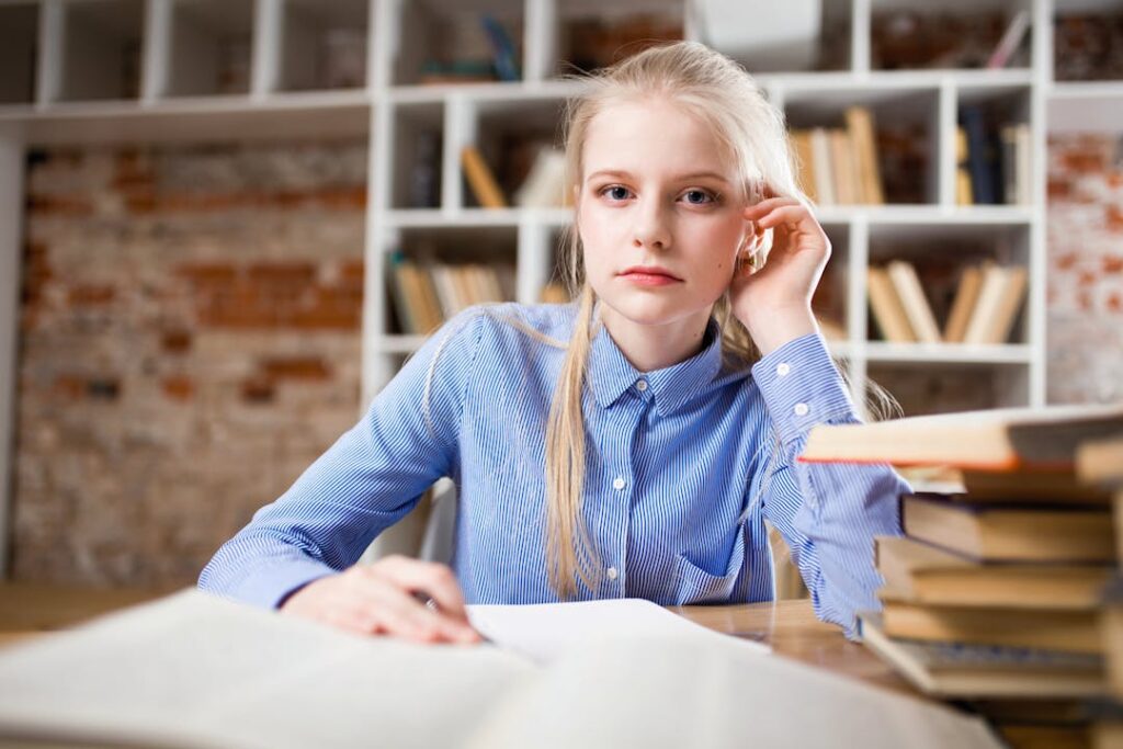 Une jolie fille en chemise bleue travaille sur un bureau avec une bibliothèque derrière.