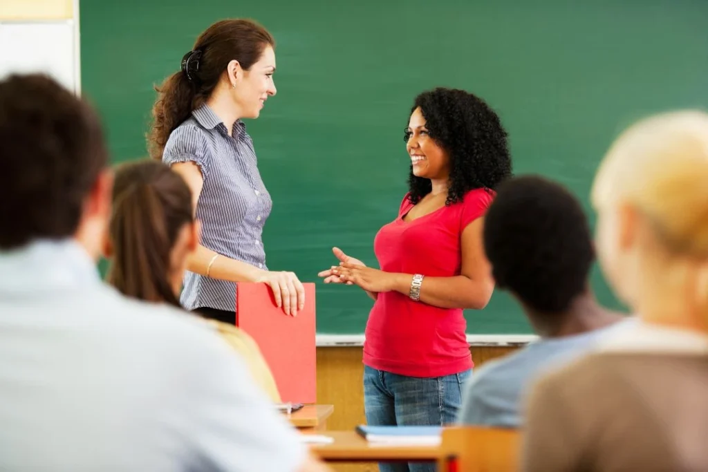 Une élève se tient debout face à sa prof, au tableau d'une salle de cours.
