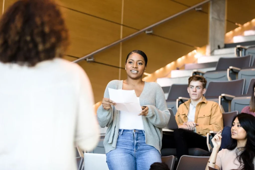 Une jeune femme se tient debout et parle devant d'autres étudiants, face à l'enseignante.