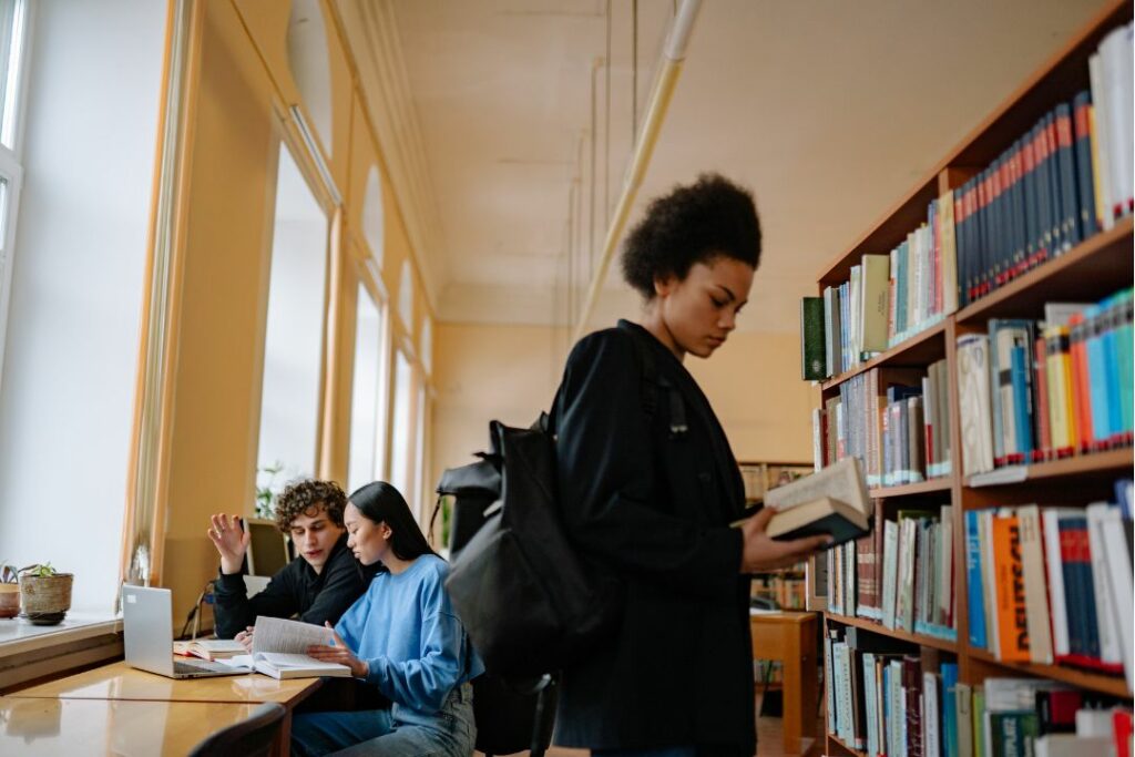 Étudiante lisant un livre debout dans une bibliothèque universitaire.