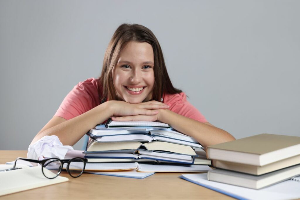 Lycéenne souriante posant le menton sur une pile de dossiers scolaires sur son bureau.
